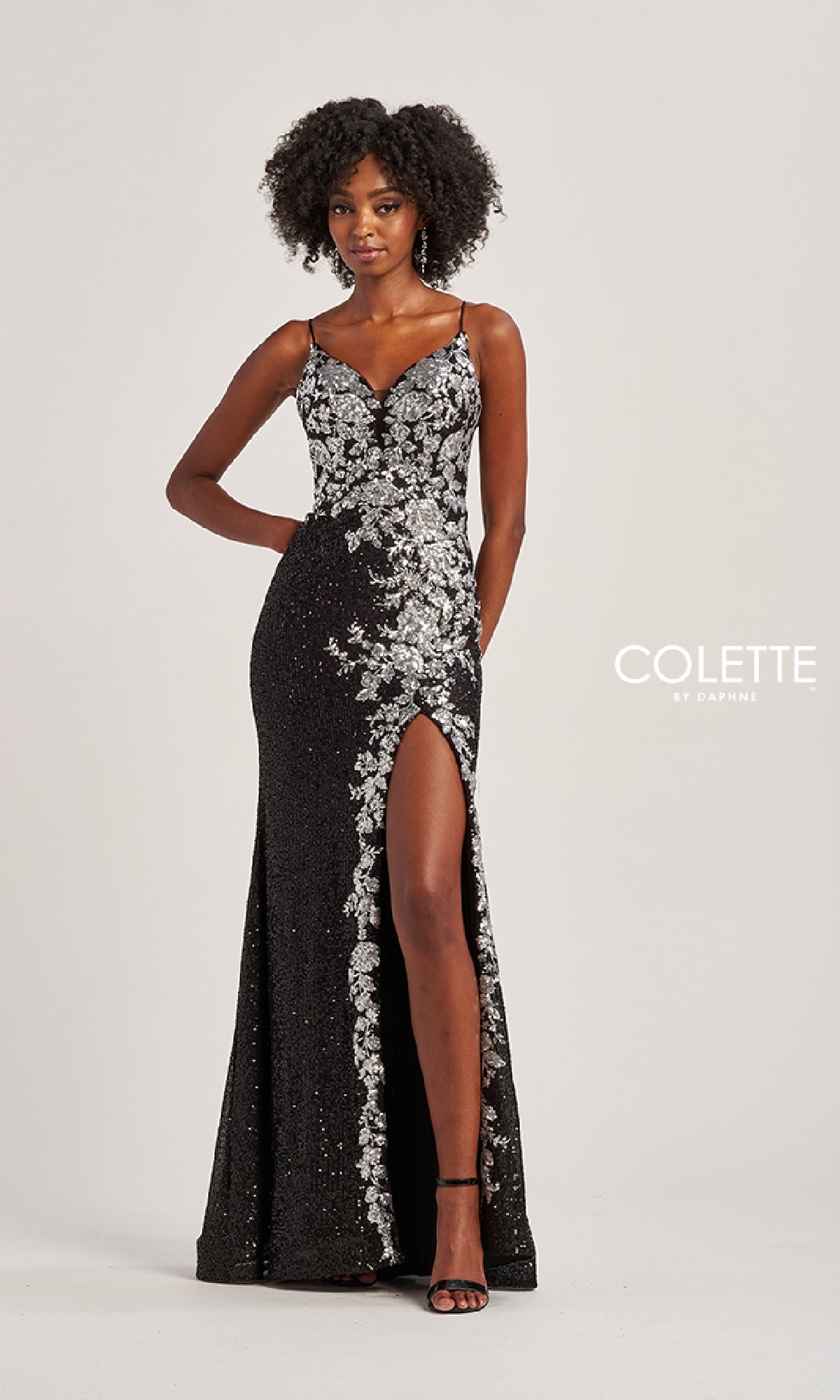 A woman models the Long Prom Dress: Colette CL8425—a black, floor-length gown with a corset bodice, silver floral sequin applique, and a front thigh-high slit—posing against a plain background.